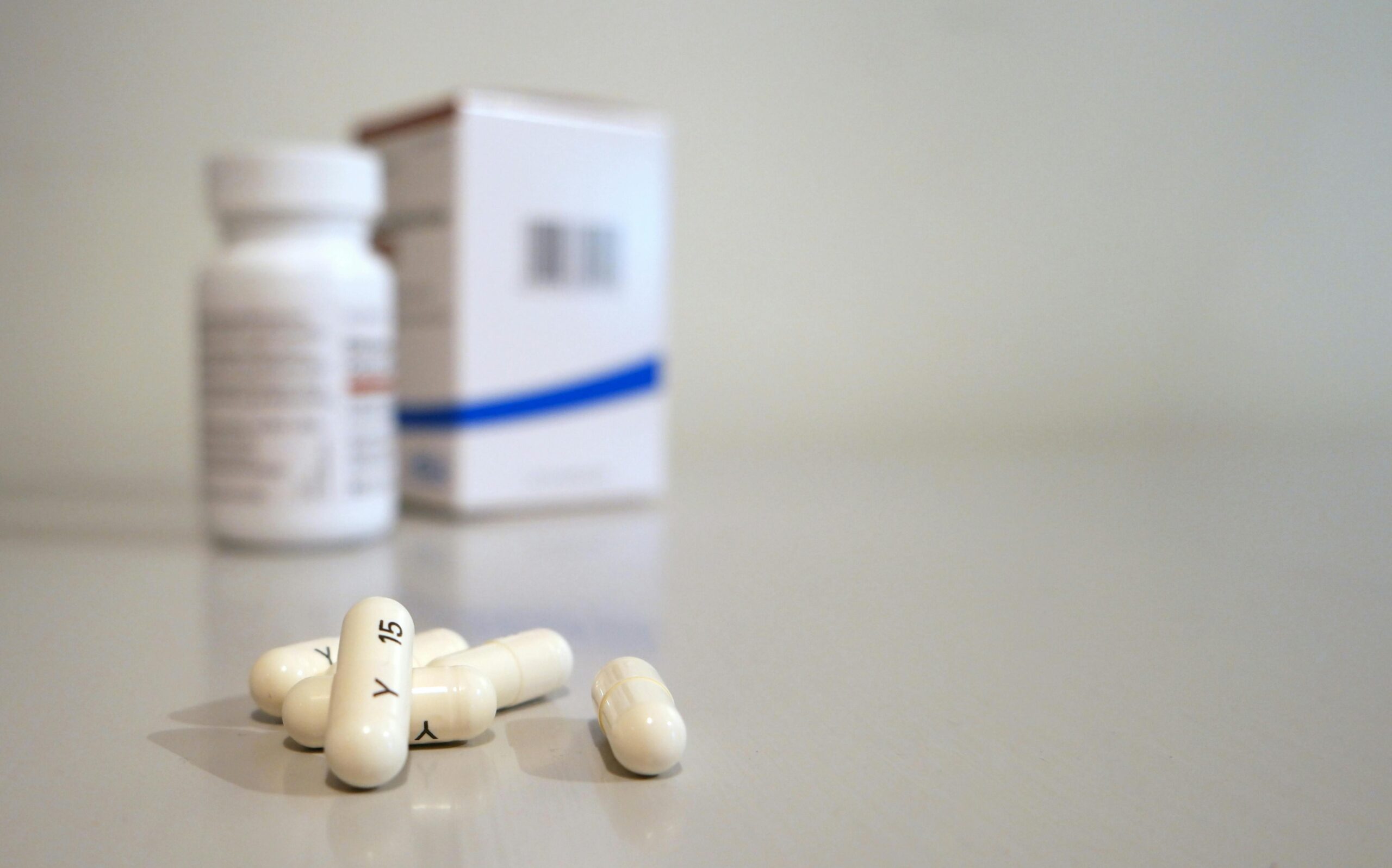 Close-up image of white capsules with medicine bottle and box in the background.
